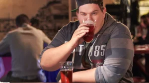 Image of stout-looking male in a rugby shirt, drinking a glass of beer with another beer in front of him waiting to be consumed - How Long Does Alcohol Stop Fat Burning - South Shores Recovery
