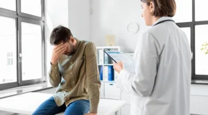 Image of a male patient sitting on a table with his hand on his forehead, talking to medical counselor - Do Mental Health Leave California Laws Cover Rehab - South Shores Recovery
